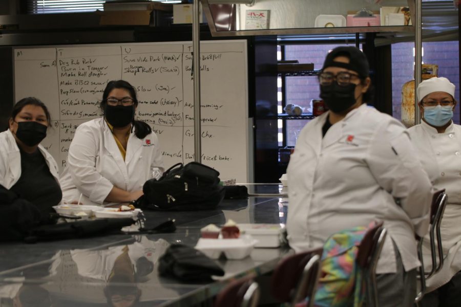 Students in the food service management 38 class pose for a photo. Behind them is a white board with a check list and each students’ role for the day. 
