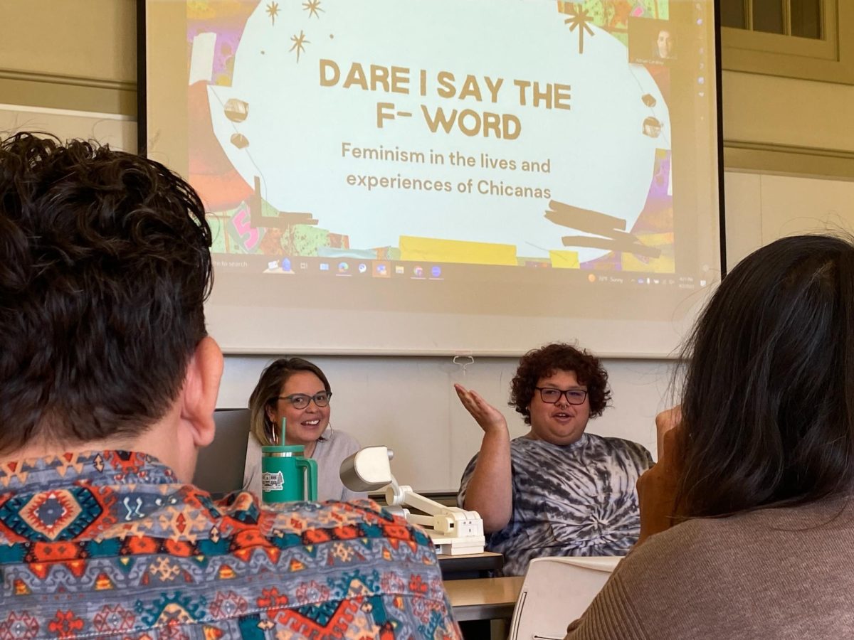 Victoria Benavides (left) and Noah Sanchez (right) introduce each other underneath a projection screen that displays the first slide of a slideshow on Chicana Feminism throughout history on April 2, 2024. 