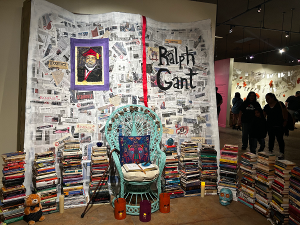 Alexandria Ramos O'Casey’s artistic ofrenda in honor of late Fresno City College professor Ralph Gant is displayed in the Arte Américas art exhibit on Oct. 2. The ofrenda is a large papier-mâché sculpture shaped to look like a book and made up of Rampage newspapers pasted on top, with Ralph Gant’s name written in black letters on the right side, and Gant’s painted portrait on the left. In front of the book structure is a teal chair with an open book in the seat, surrounded by stacks of Gant’s personal books and candles. 